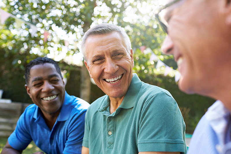 Three older men smiling and talking together outdoors in a sunny backyard.