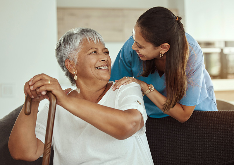 Home care worker smiling and talking with an older woman holding a cane in a living room.