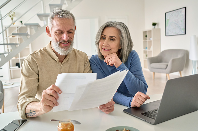 Older couple reviewing paperwork together at a table with a laptop in a bright home.