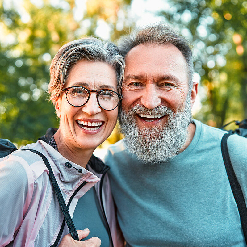 Smiling older couple outdoors wearing backpacks, posing for a close-up selfie in a park.