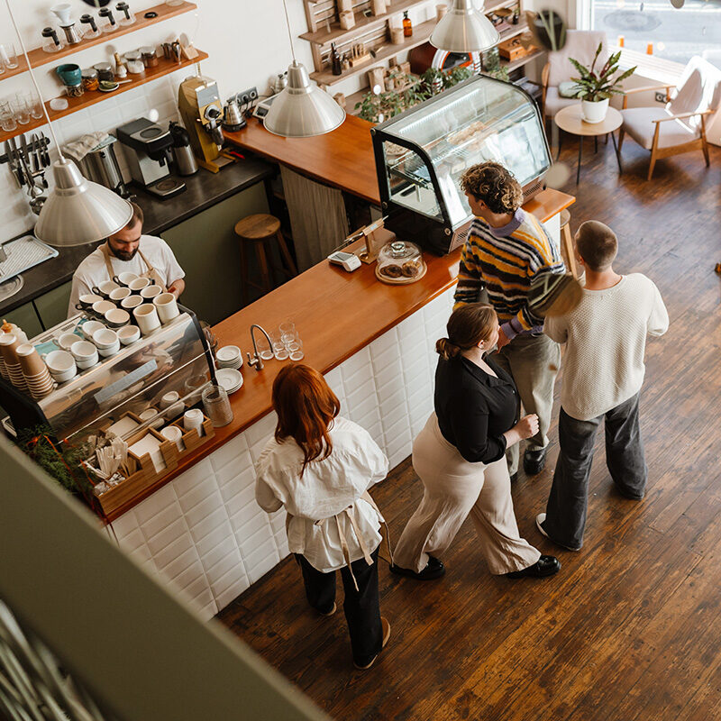 Overhead view inside a coffee shop showing a barista behind the counter and several customers waiting near a pastry display on a wood floor.