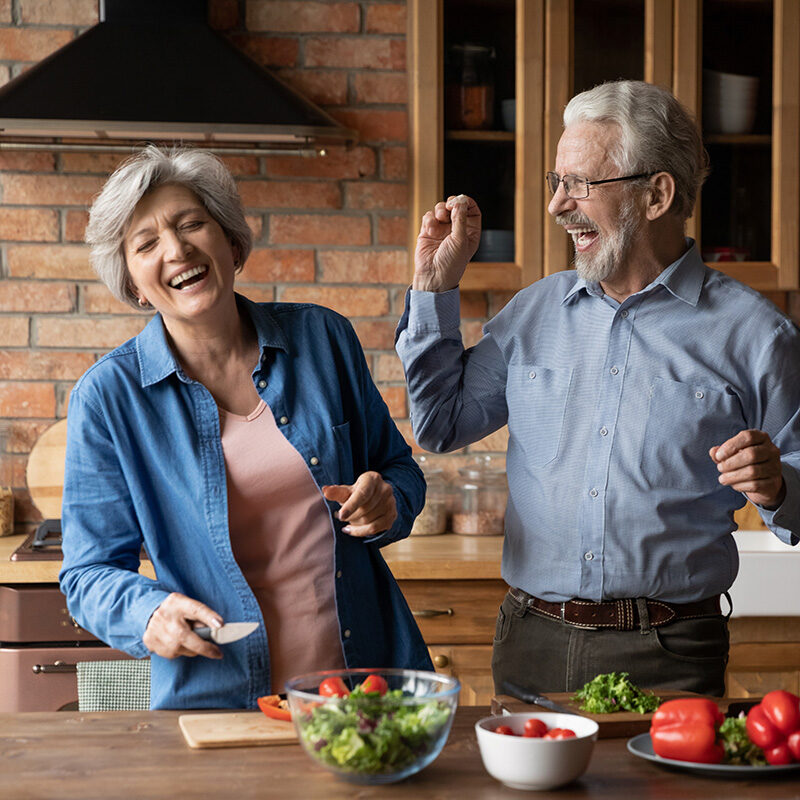 Older couple laughing together while preparing a salad in their kitchen.