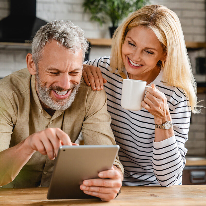 Smiling middle-aged couple at home looking at a tablet together while the woman holds a coffee mug.